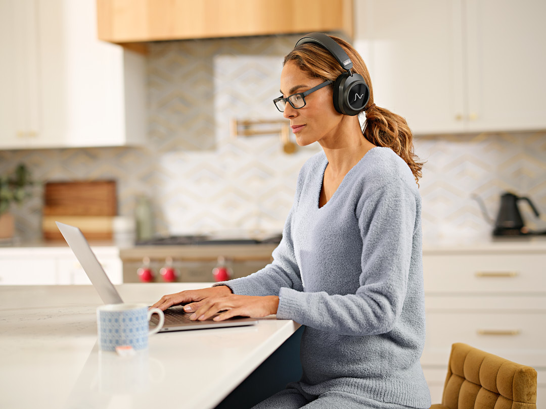 A woman working at her laptop in the kitchen, wearing Neurable One headphones