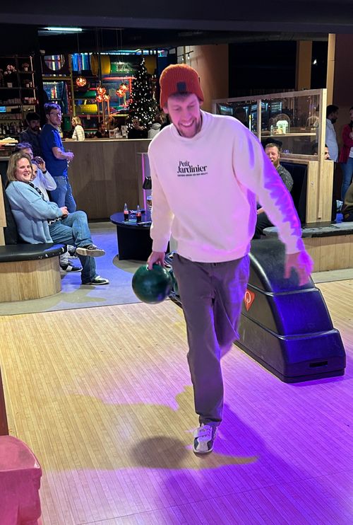 Man met oranje muts die een groene bowlingbal vasthoudt op een bowlingbaan, terwijl mensen op de achtergrond toekijken.