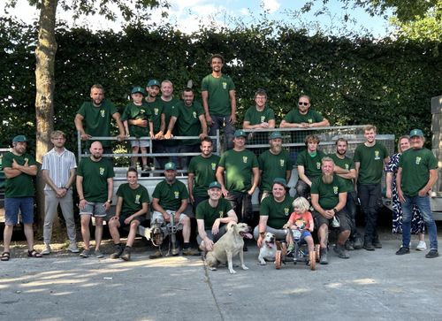 Groep van volwassenen en kinderen in groene teamshirts poseren buiten met twee honden en een driewieler-kind.