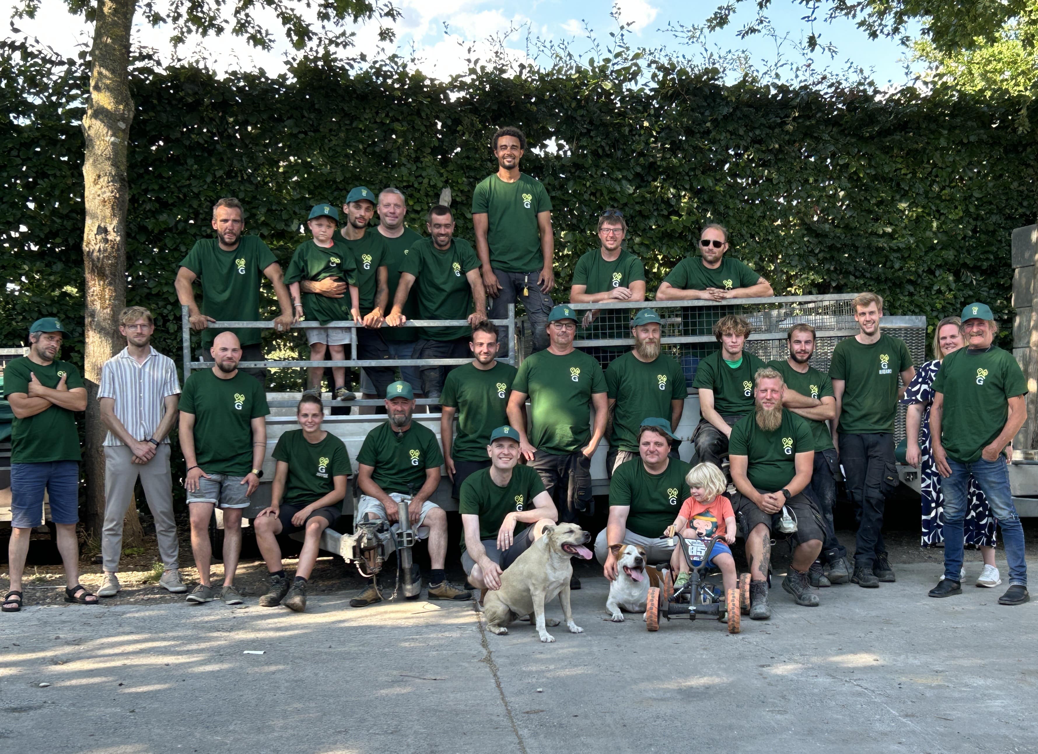 Groep van volwassenen en kinderen in groene teamshirts poseren buiten met twee honden en een driewieler-kind.