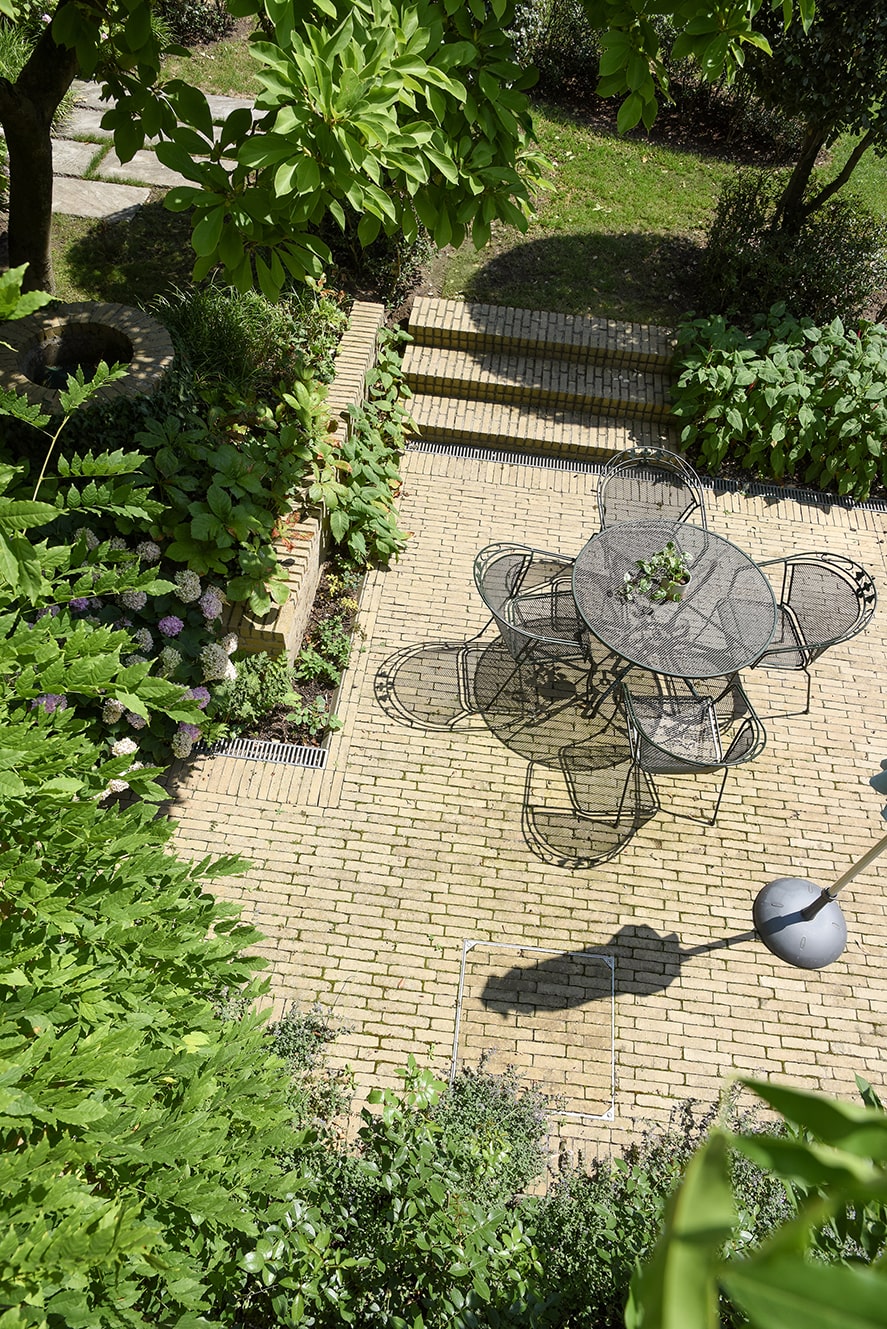 Bovenaanzicht van een zonnig terras met een ronde metalen tafel en vier stoelen omgeven door groen en planten.