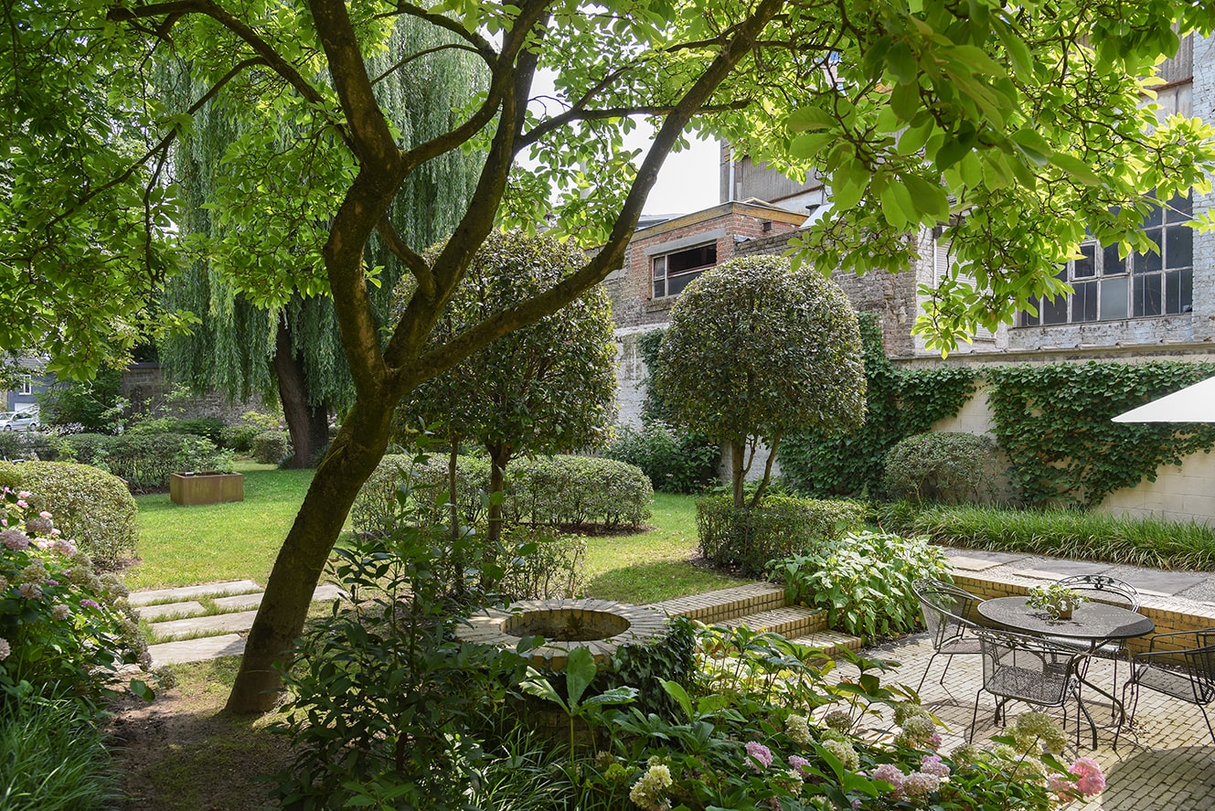 Gezellige stedelijke tuin met groene bomen, struiken en een patio met zwart metalen tafel en stoelen.