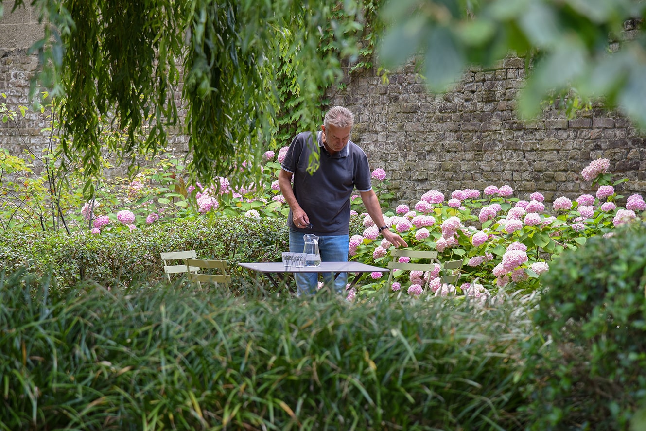Man die een groene stoel bij een tafel met glazen en een karaf in een tuin met roze hortensia's en een stenen muur achter hem zet.