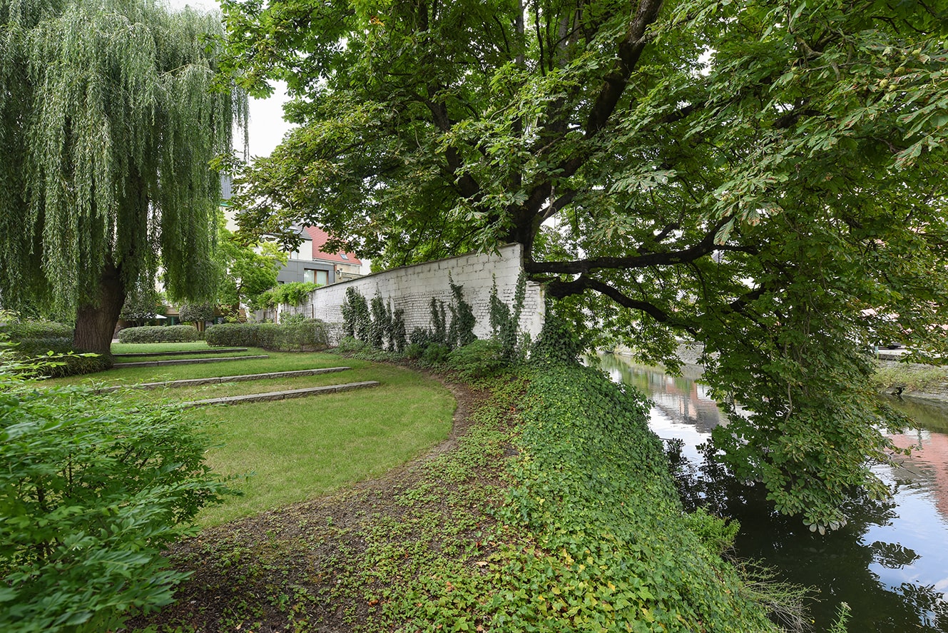 Groene tuin met gras, een witte bakstenen muur met klimop, grote bomen en een waterloop aan de rechterkant.