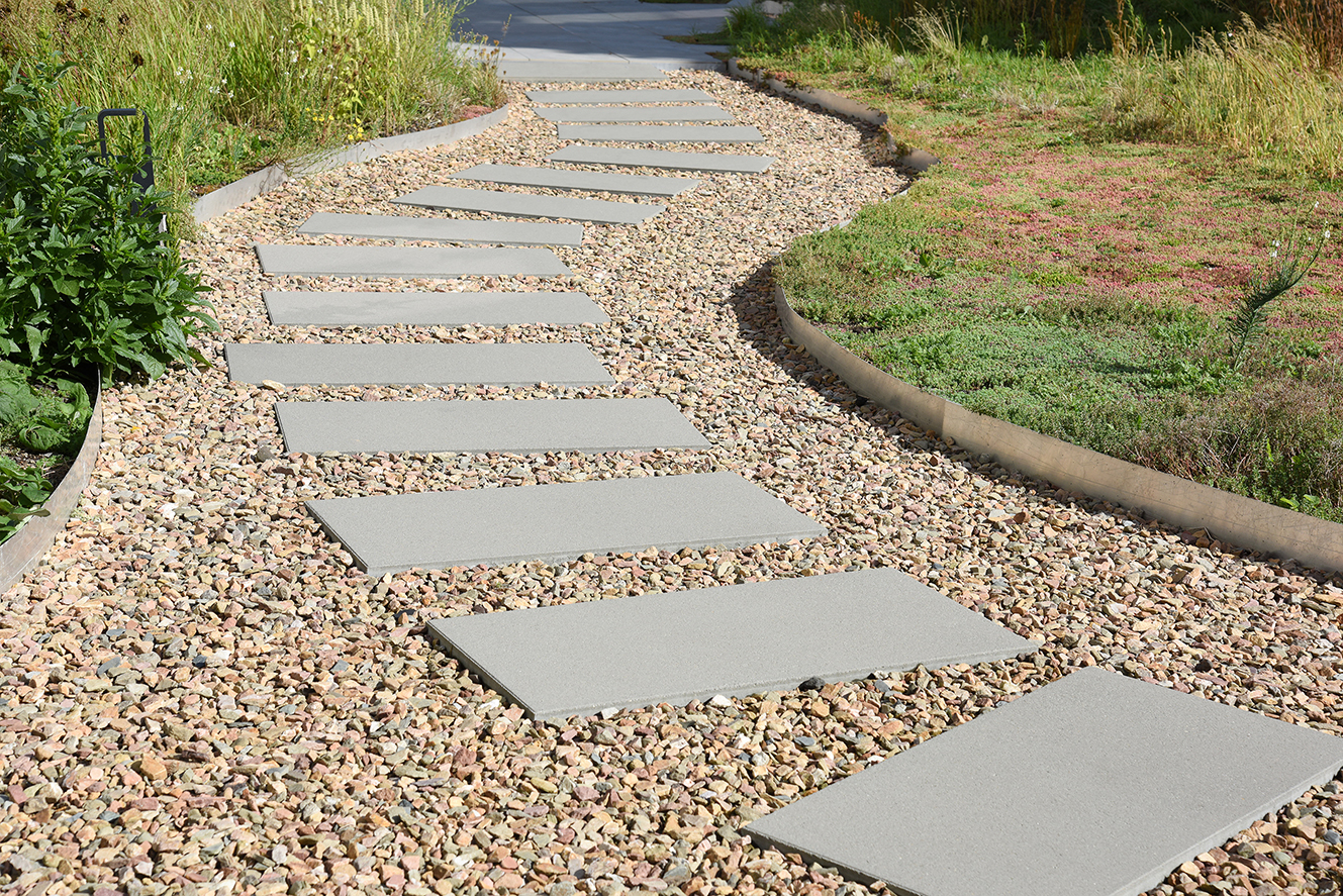 Sierpad met grote, rechthoekige betonnen tegels op kleine kiezelstenen, begrensd door groene planten en gras.