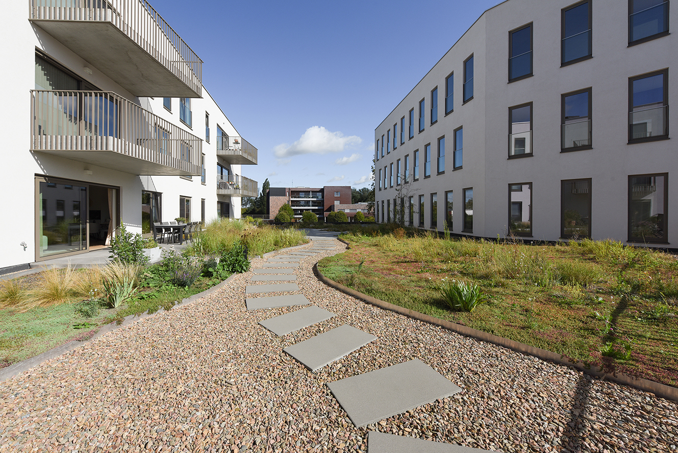 Steenpad met grote tegels tussen twee moderne witte gebouwen met balkons en planten onder een blauwe lucht.