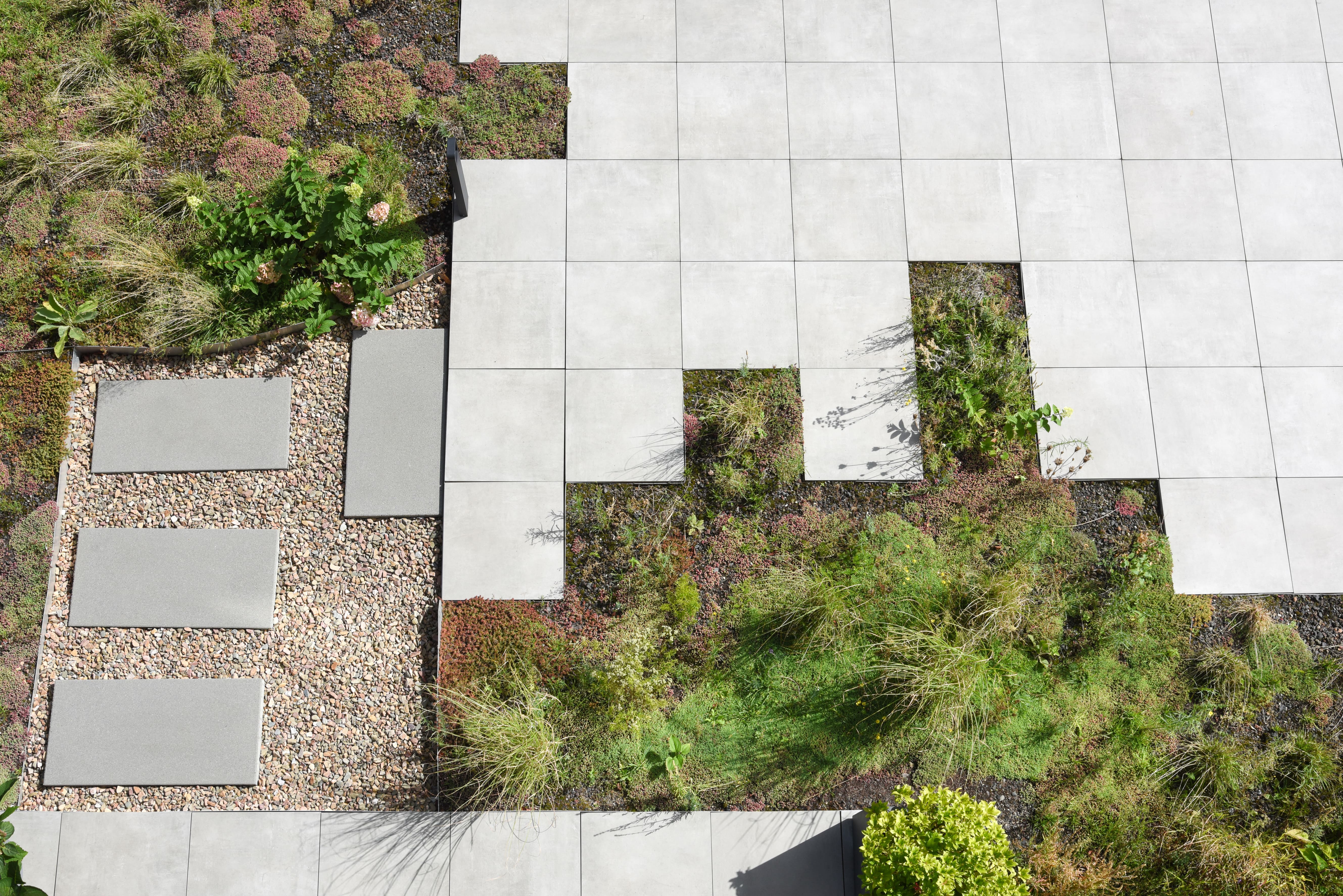 Luchtfoto van een tuin met betonnen tegels, grindpad met drie betonnen platen en diverse groene planten en gras.