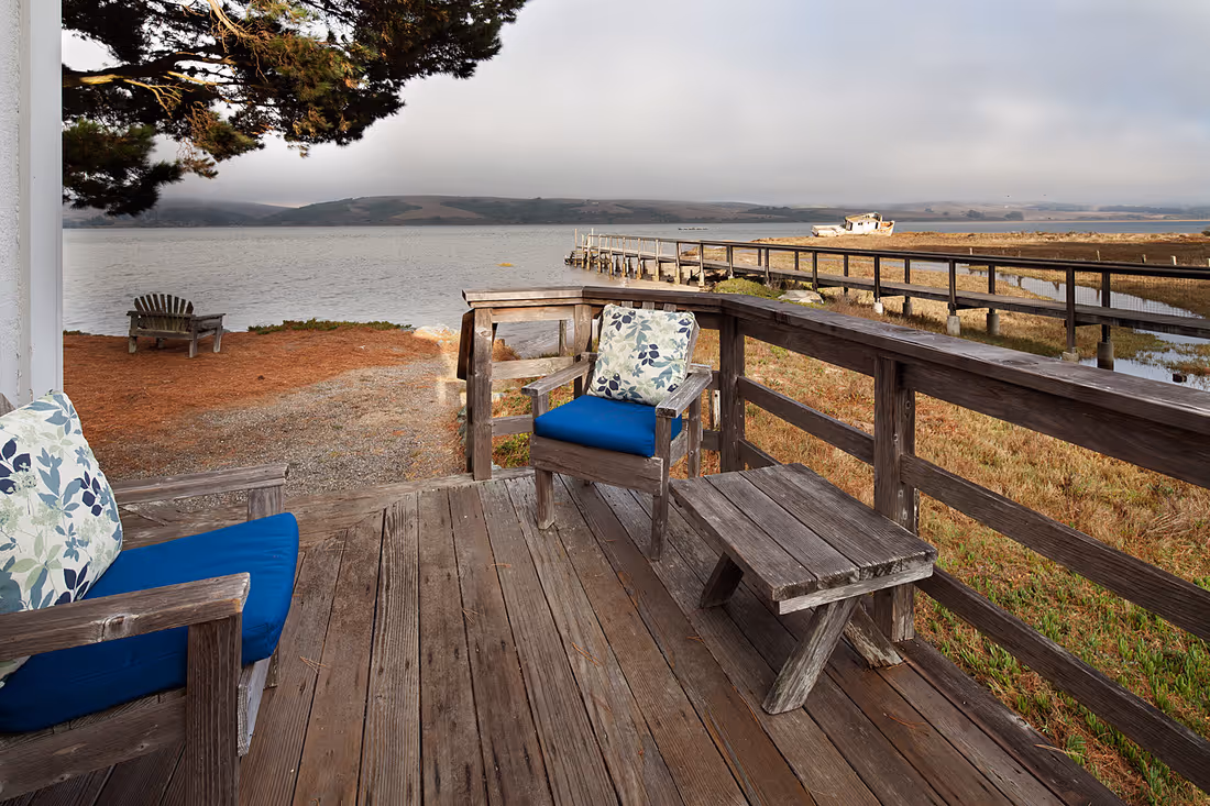 Wooden deck with cushioned chairs overlooking a calm body of water and a dock extending into the water on a cloudy day.