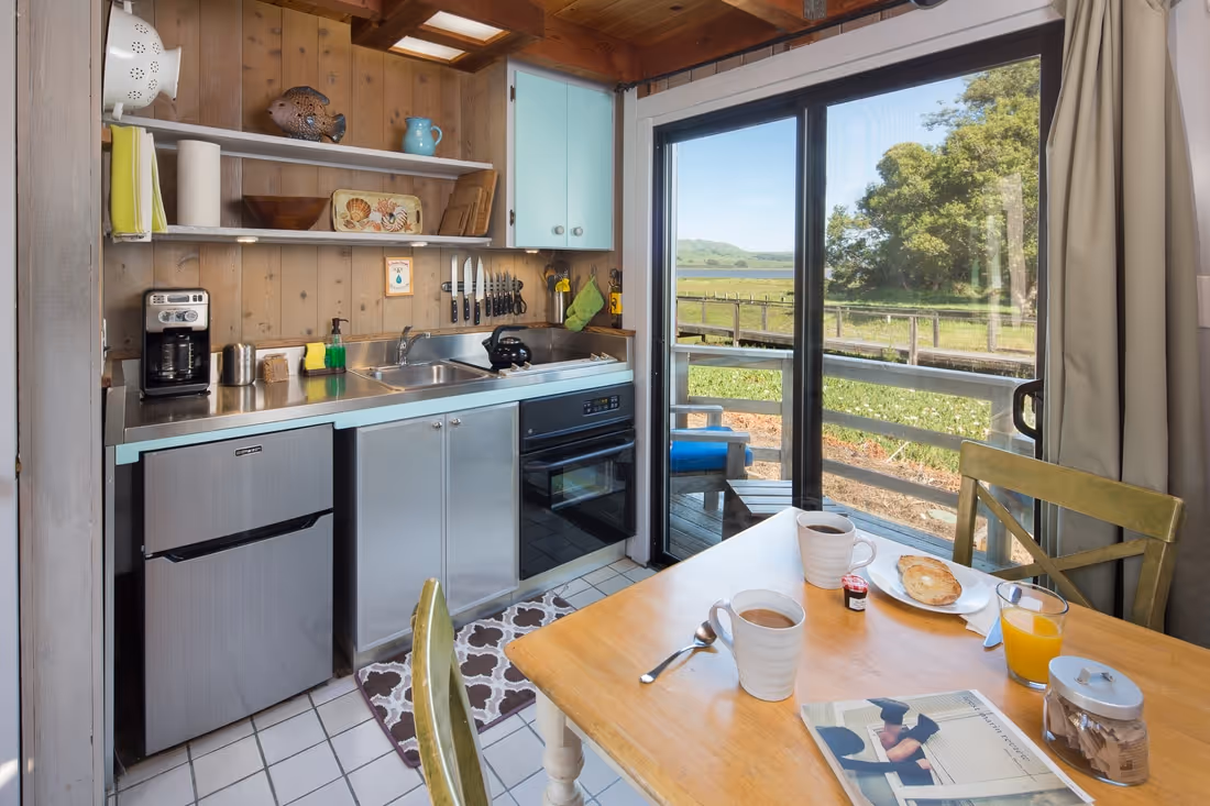 Compact kitchen with stainless steel appliances, wooden walls, and a table set for breakfast by a sliding glass door overlooking a green outdoor area.