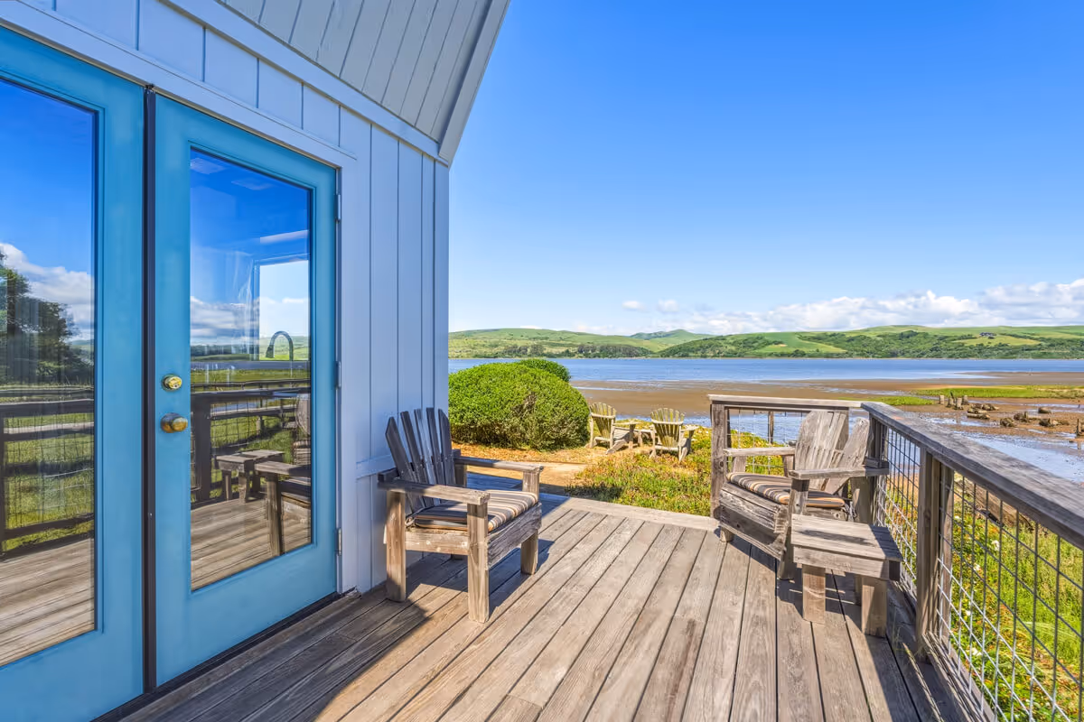 Wooden deck with chairs overlooking a calm body of water, green hills in the distance, and clear blue sky.