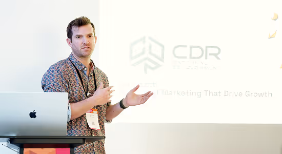 Brooke giving a presentation in front of a screen displaying a company logo and text, standing beside a laptop on a podium.