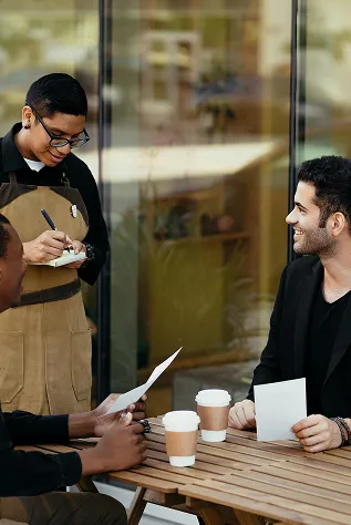 male waiter taking an order for two male patrons sat at a table with two coffees