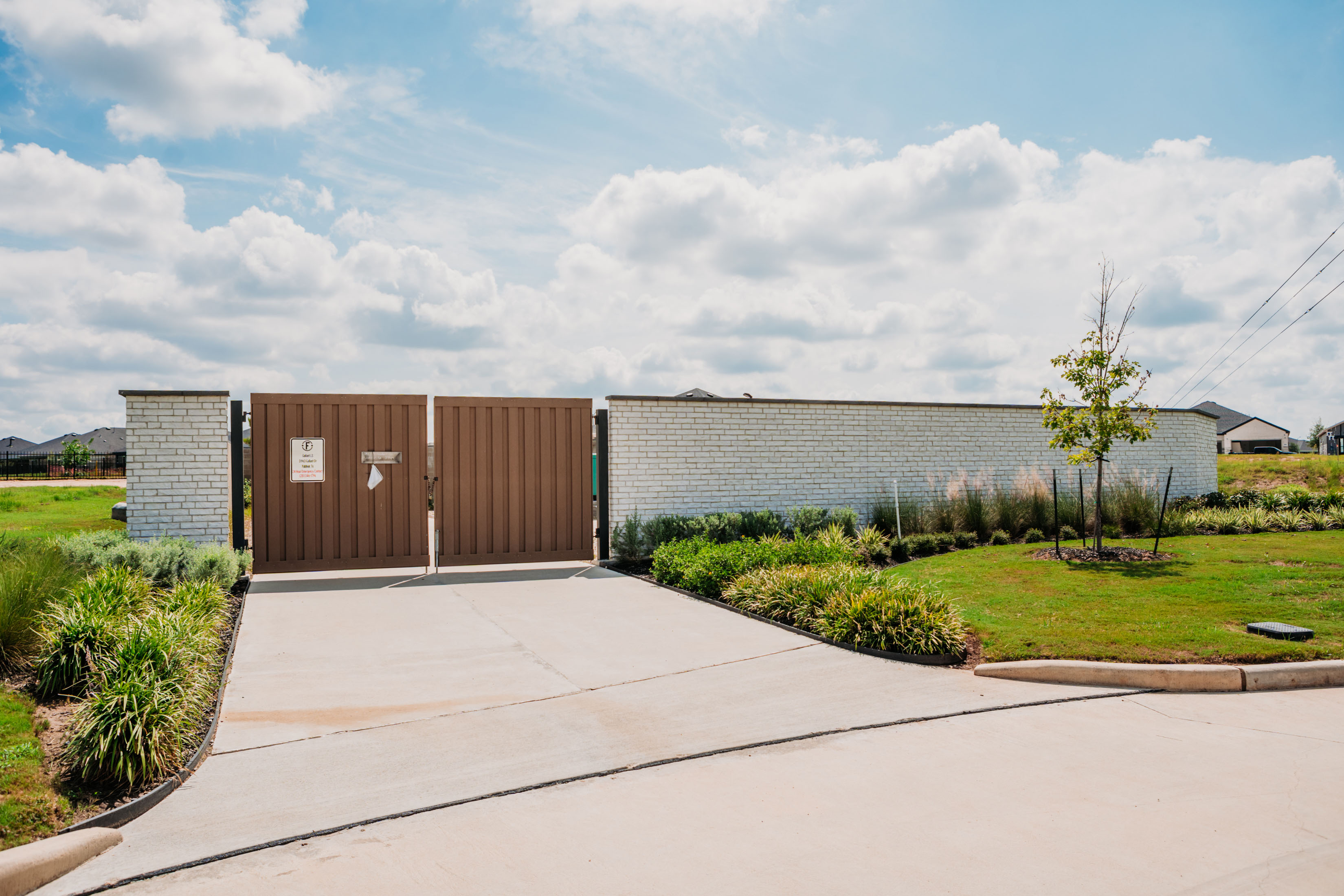 A community entrance featuring a white brick wall with pilasters flanking a wide brown wood-paneled gate and a paved driveway, surrounded by green landscaping under a partly cloudy sky.