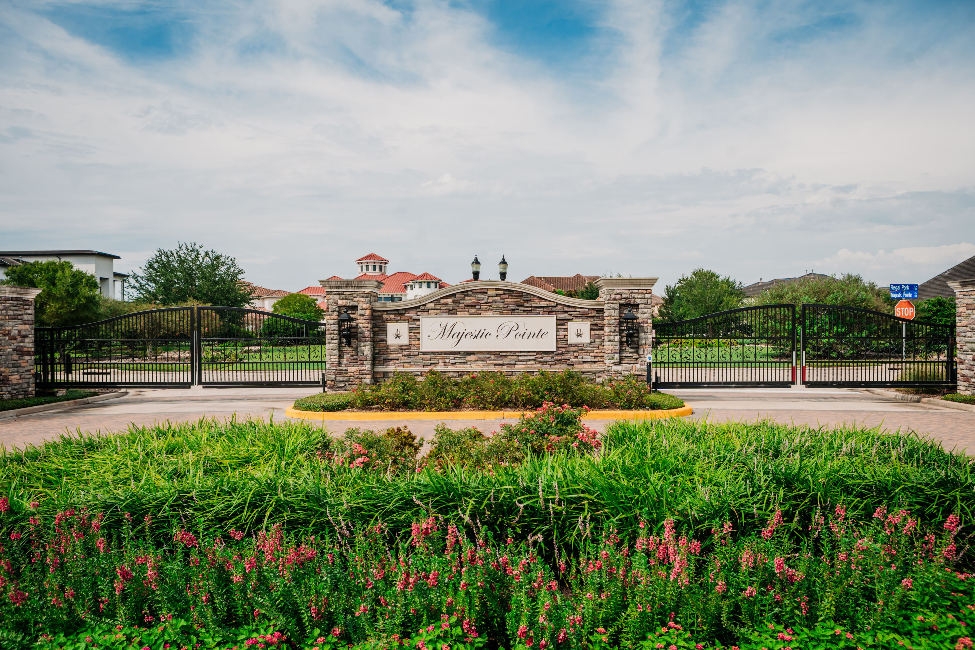 The grand entrance to "Majestic Pointe," featuring a central stone and stucco monument sign flanked by stone pillars and two wide black wrought iron gates, surrounded by lush green and pink flowers.