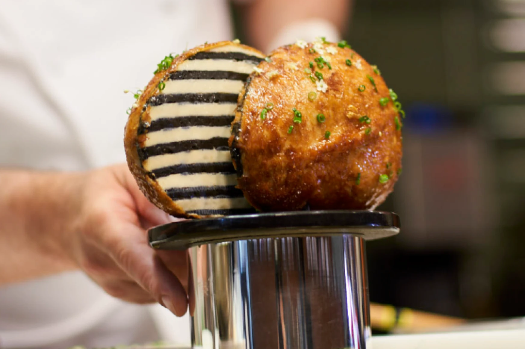 Close-up of a hand holding a gourmet dish with a circular fried pastry topped with chopped herbs and a striped black and white filling inside.