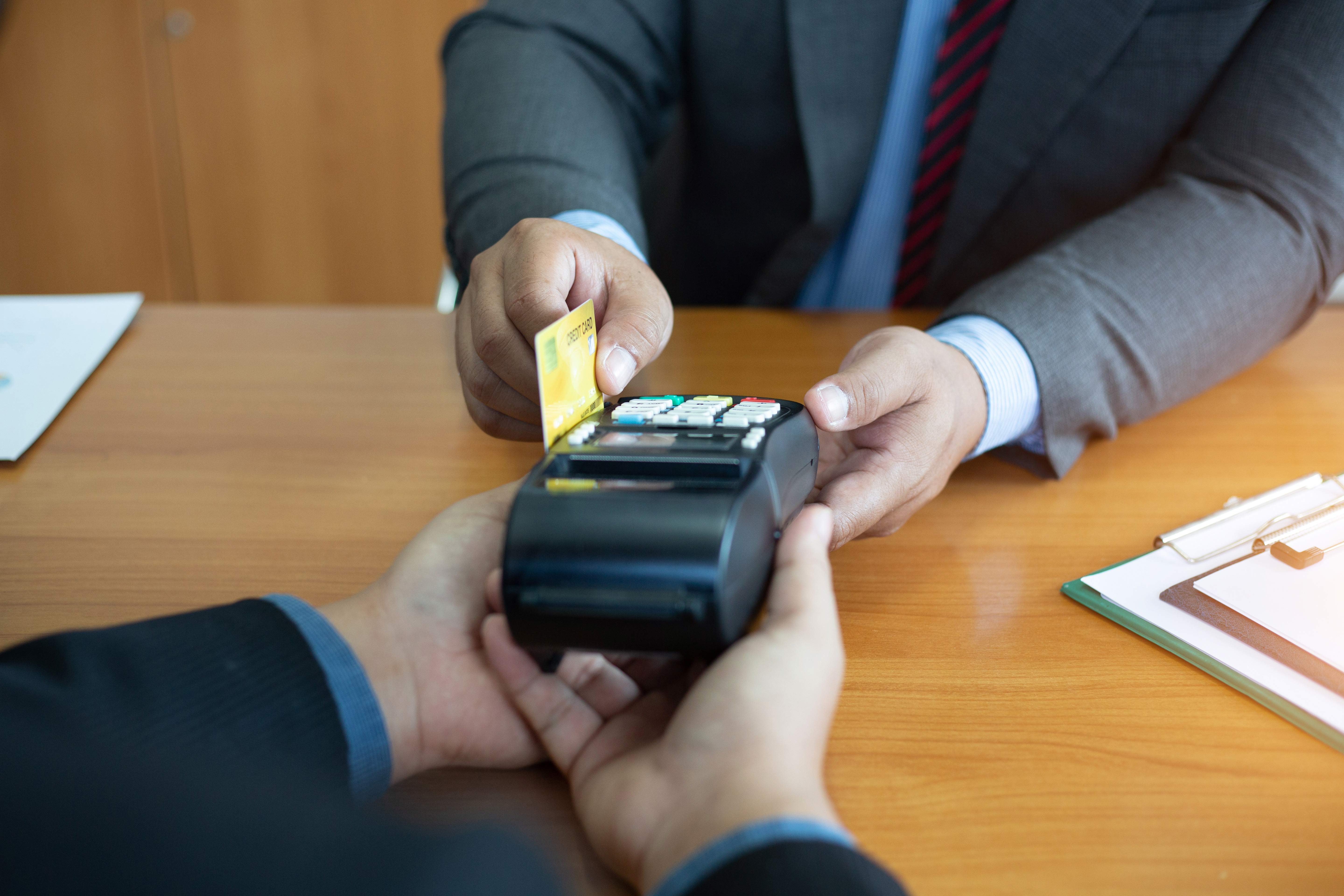 Stressed man sitting at a table with a laptop, coffee cup, and open planner while a woman and child play in the background of a modern living room.