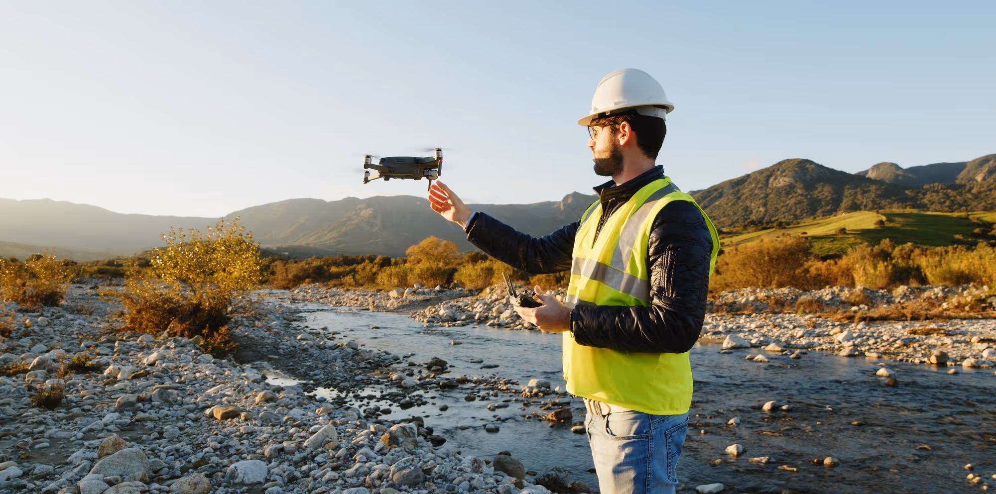 Man wearing a white construction helmet and yellow safety vest operating a drone near a rocky river with mountains in the background.