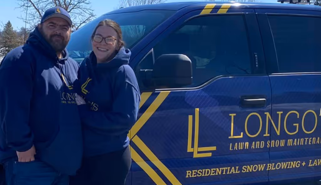 Smiling man and woman wearing matching Longo's Lawn and Snow Maintenance hoodies standing next to a blue company van with yellow logo and text.