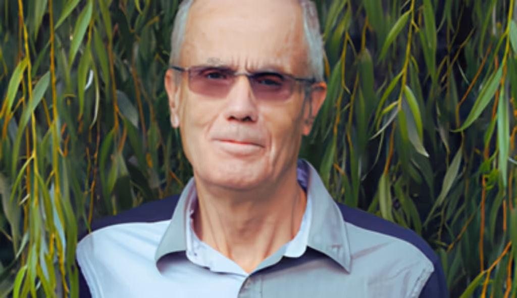 Older man wearing sunglasses and a collared shirt standing in front of green leafy plants.