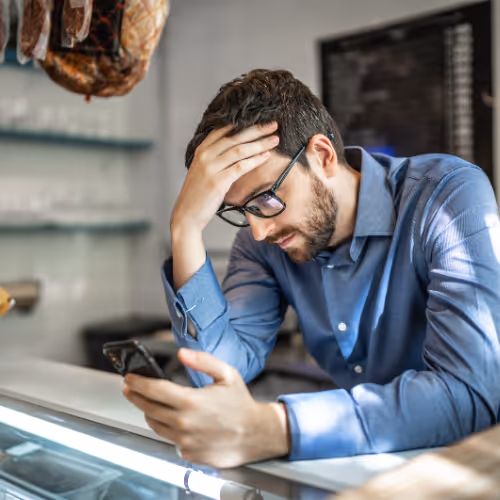A man sitting at his work counter looking at his phone with disappointment on his face. 