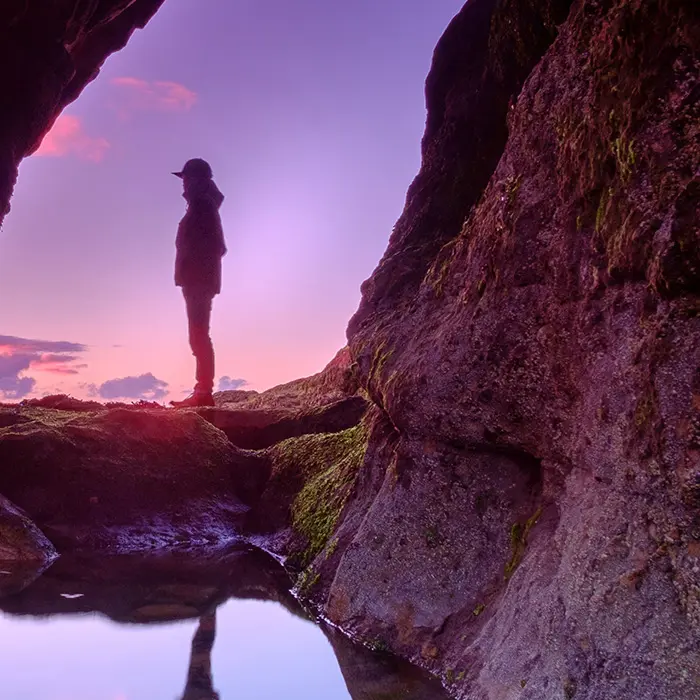 Person standing between coastal rocks at sunset, reflected in a tide pool.