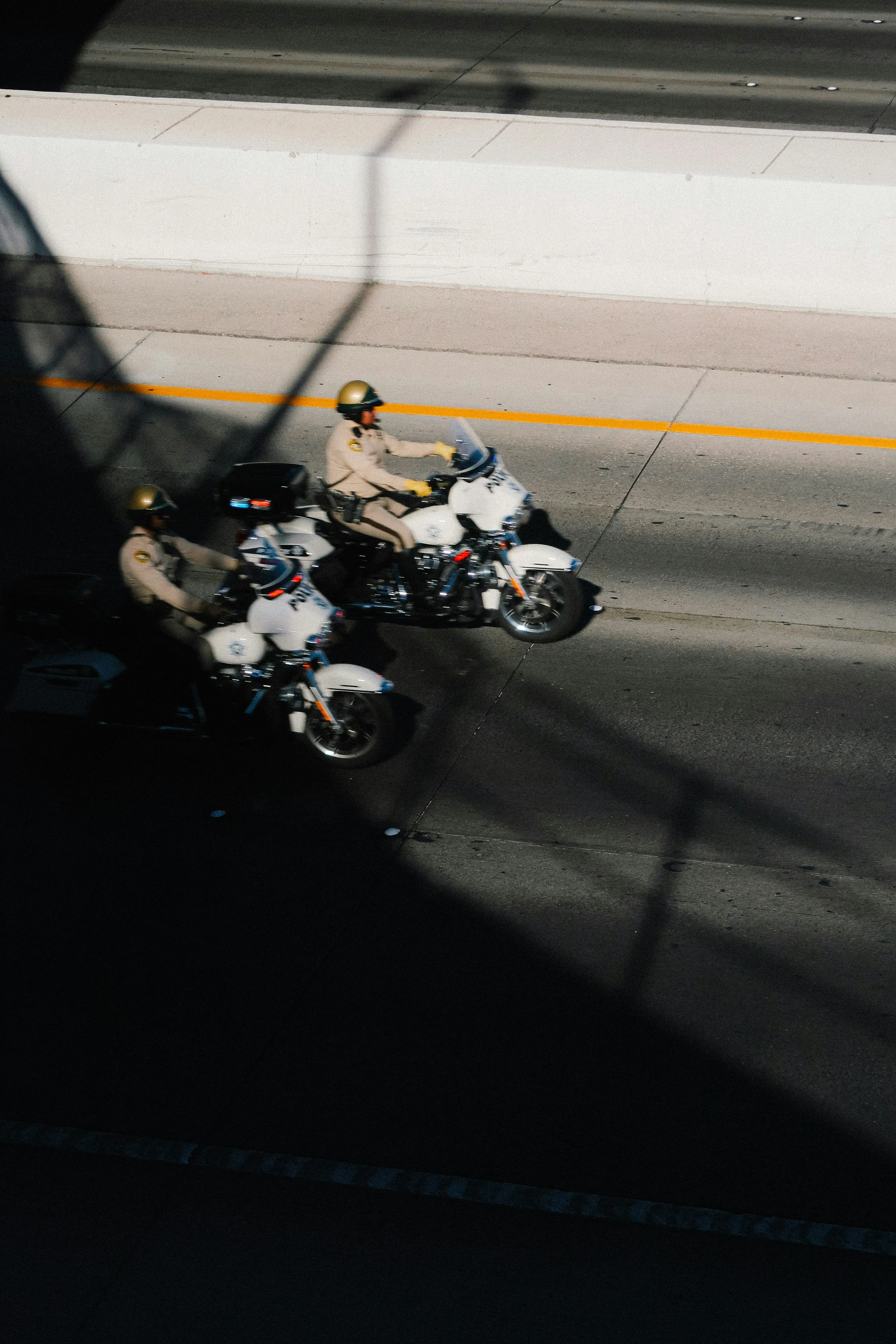 Two uniformed police officers riding motorcycles on a highway, captured from an elevated angle in bright sunlight.