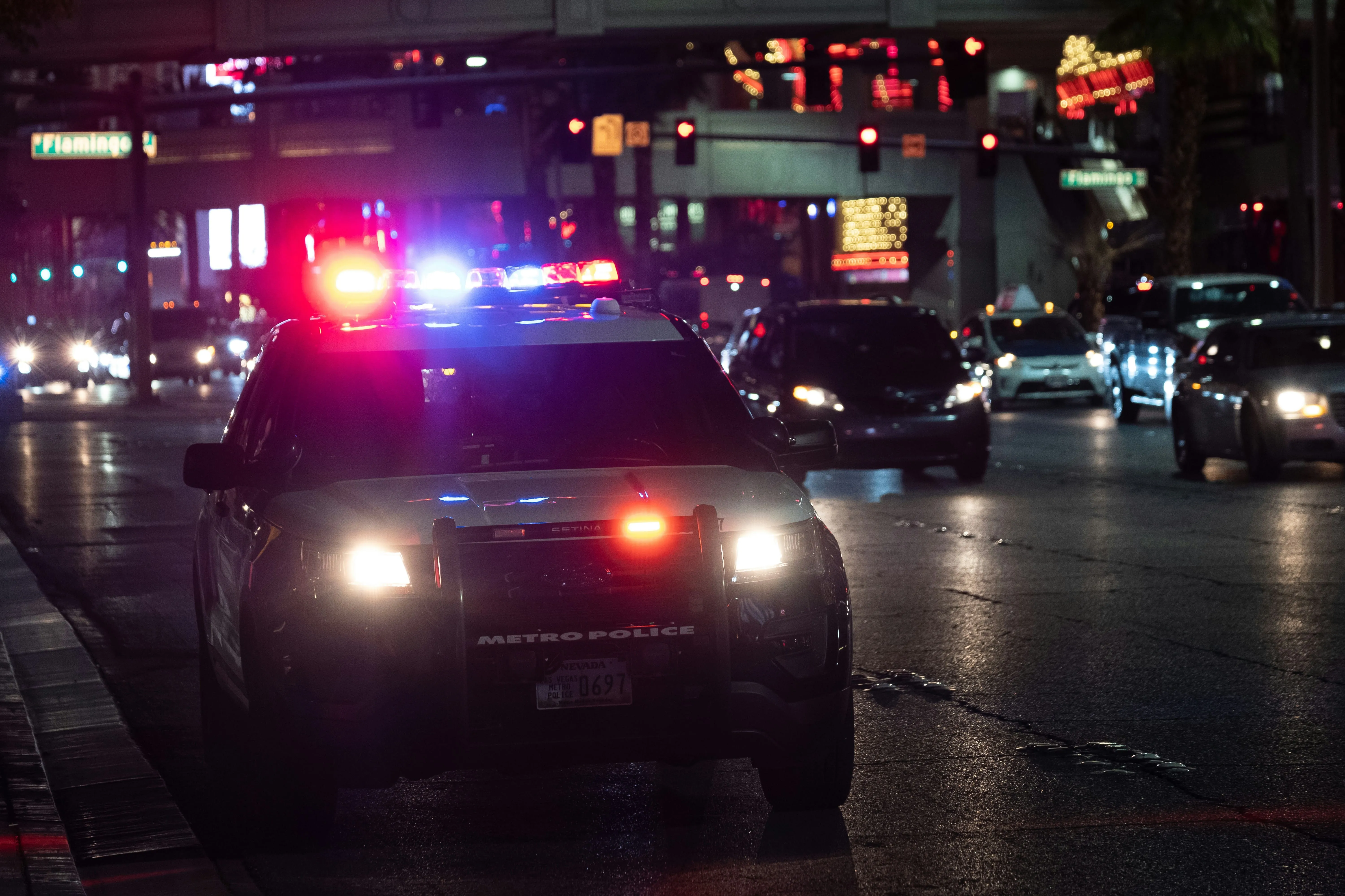 Police SUV with flashing red and blue lights parked on a busy street at night, with city traffic and lights in the background.