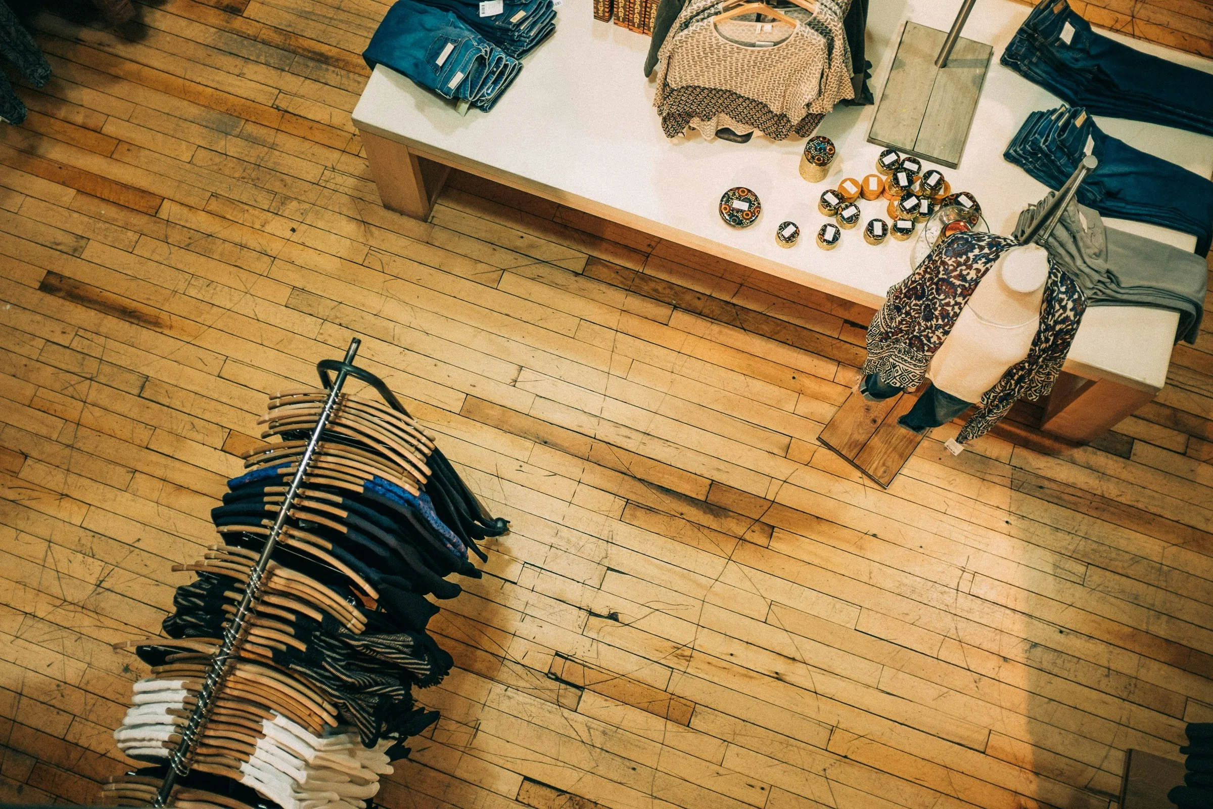 Overhead view of a boutique retail store with folded clothes, display items, and garments on hangers, laid out on a wooden floor.