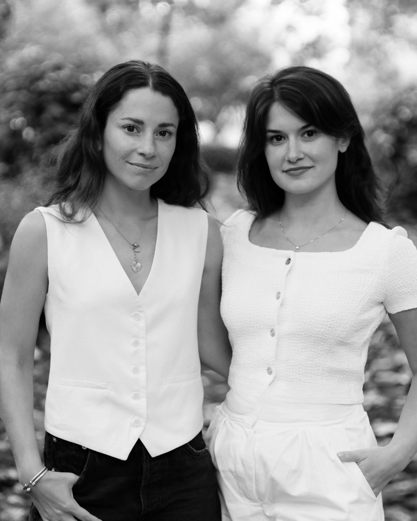 Two women standing outdoors with arms around each other, both wearing buttoned white tops and looking at the camera.