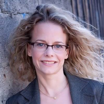 Portrait of a smiling woman with curly light brown hair wearing a grey top against a gray background. Eva Schiffer.