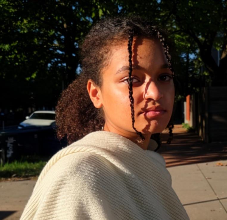 Young woman with curly hair tied back, two braided strands framing her face, wearing a light-colored textured shawl, standing outdoors on a sidewalk with trees and parked cars in the background during sunlight.