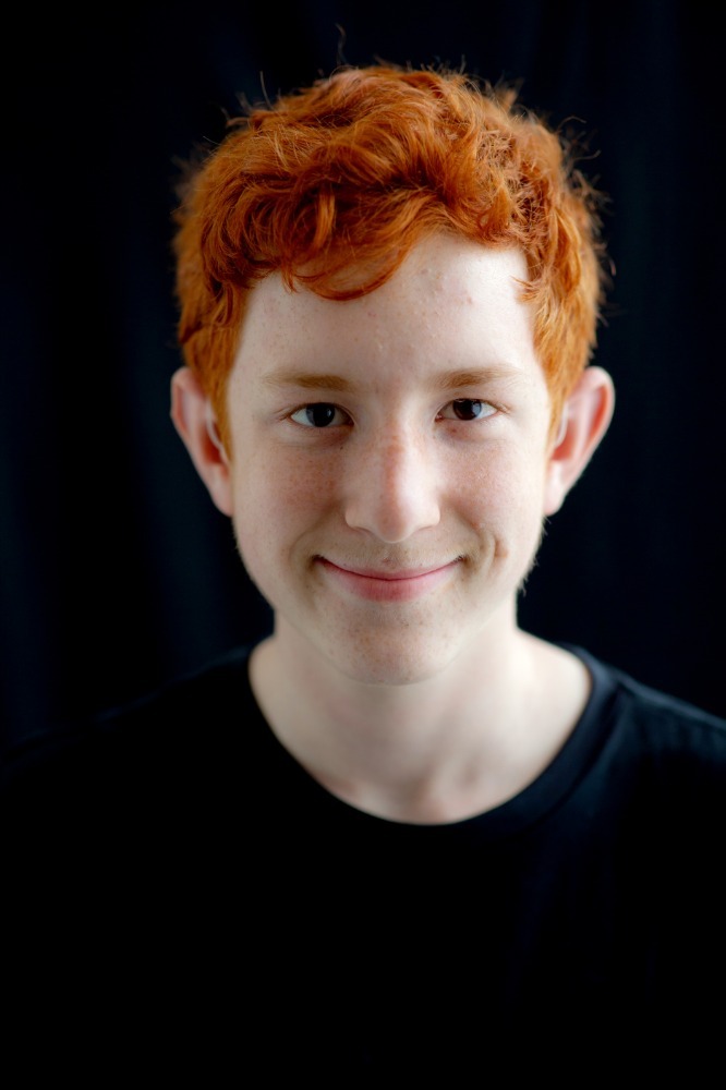 Portrait of a smiling young man with bright red curly hair and fair skin against a black background.