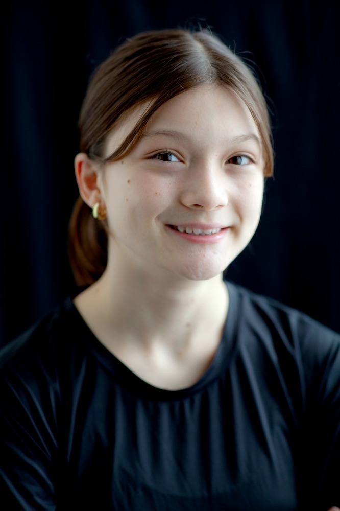 Portrait of a smiling young woman with light skin, brown hair pulled back, wearing small gold hoop earrings and a black shirt, against a dark background.