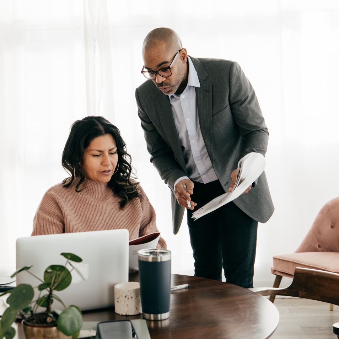A woman in a pink sweater sits at a desk with a laptop while a man in a gray suit stands beside her holding documents, both engaged in a discussion in a bright modern office.

Team collaboration in action: A business meeting between a manager and team member discussing project strategies and reviewing documents in a modern office. Boost workplace productivity with clear communication and strategic planning.