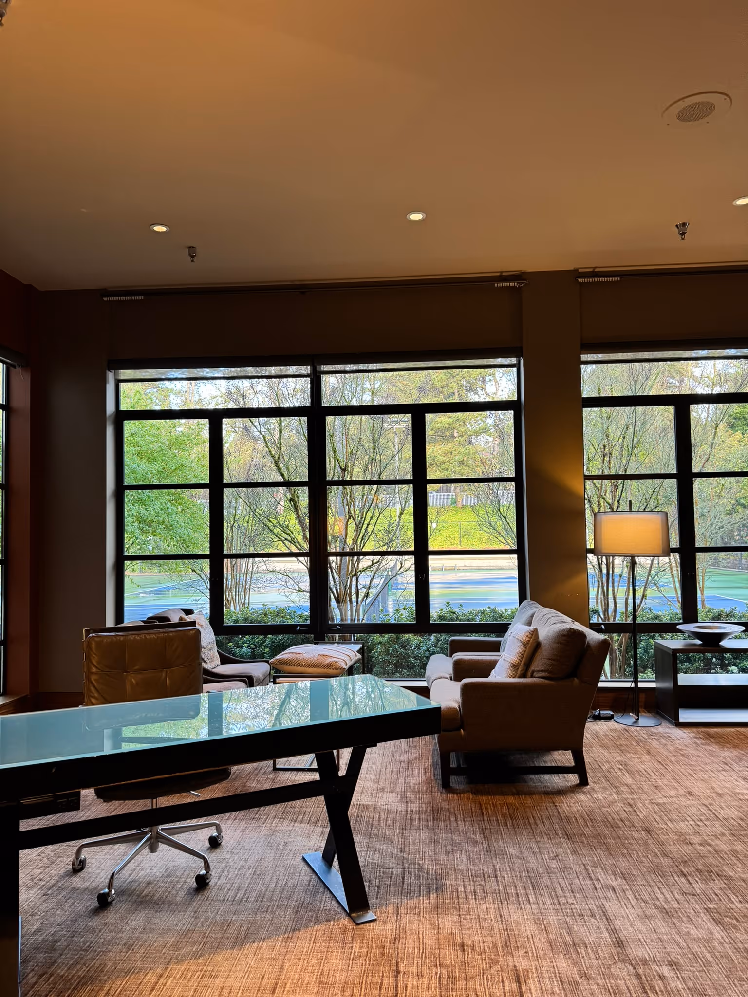 Modern office with a glass desk, rolling chair, armchairs, and large windows showing trees outside.