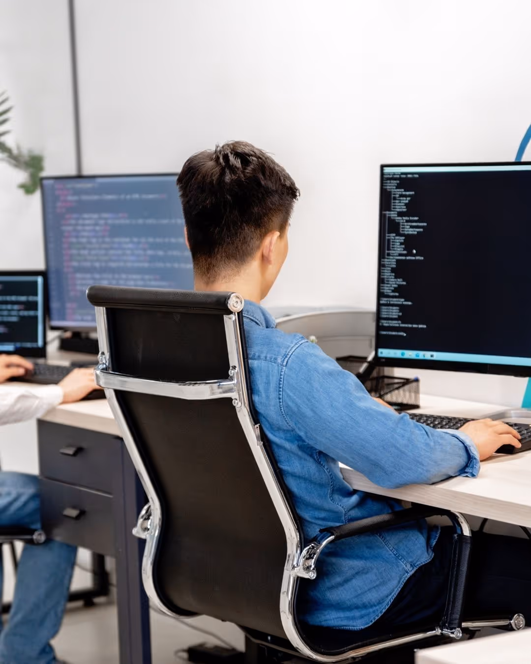 Person in a denim shirt sitting on an office chair working on a desktop computer with code displayed on the screen.