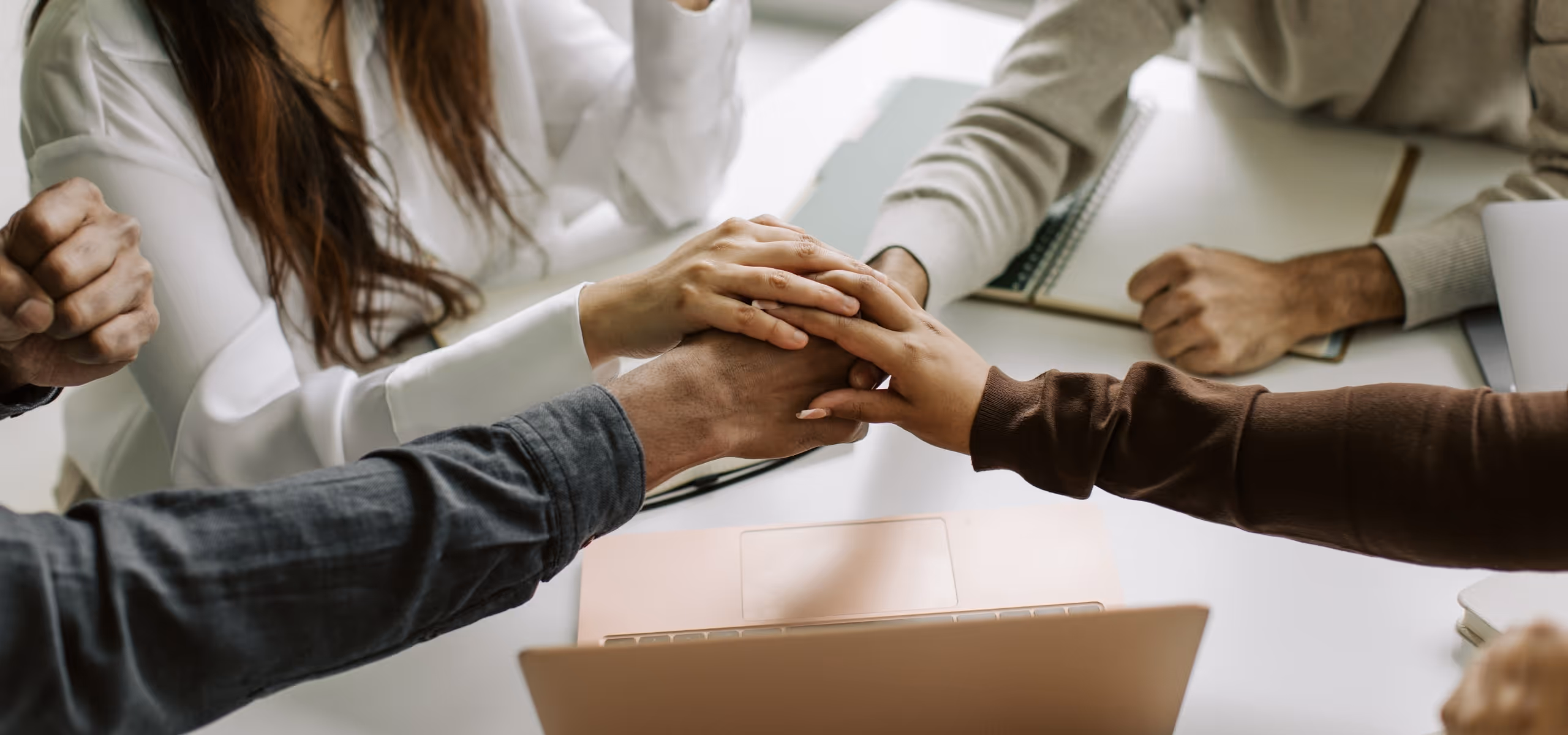 Group of people seated at a table with hands stacked together in a gesture of teamwork and support over a laptop.