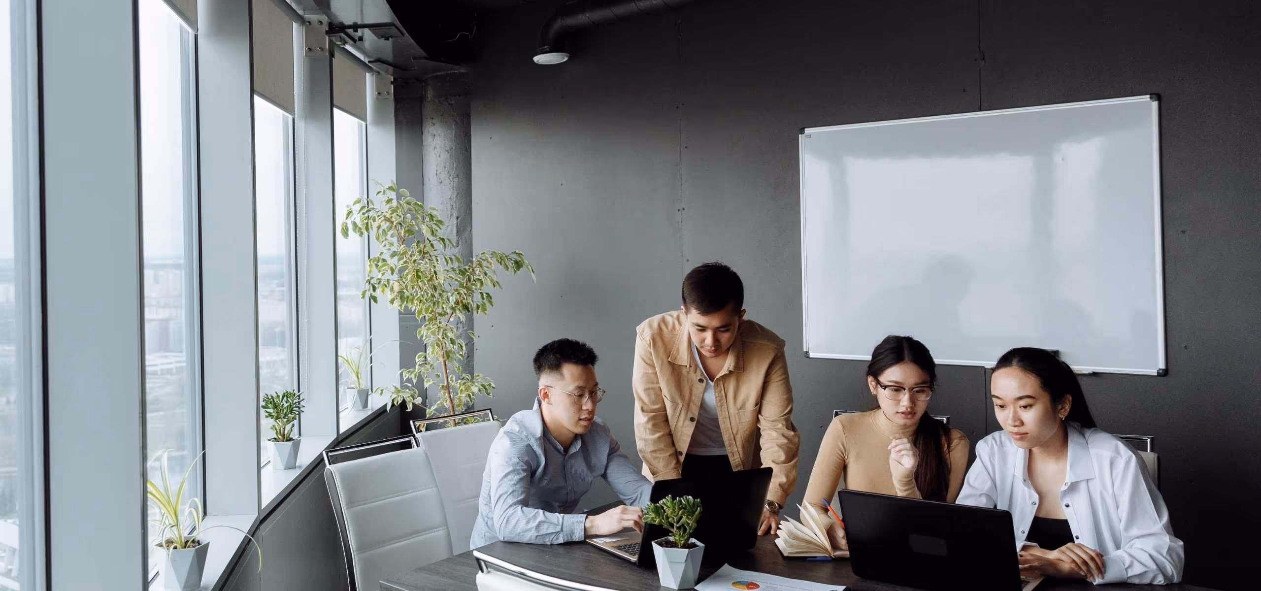 Four young professionals collaborating in a modern office with laptops, notebooks, and large windows.
