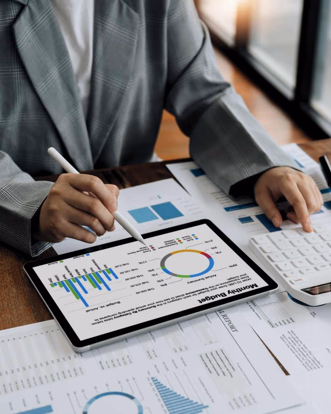 Person using a stylus on a tablet displaying a monthly budget chart with a pie chart and bar graph, next to a calculator and financial documents.