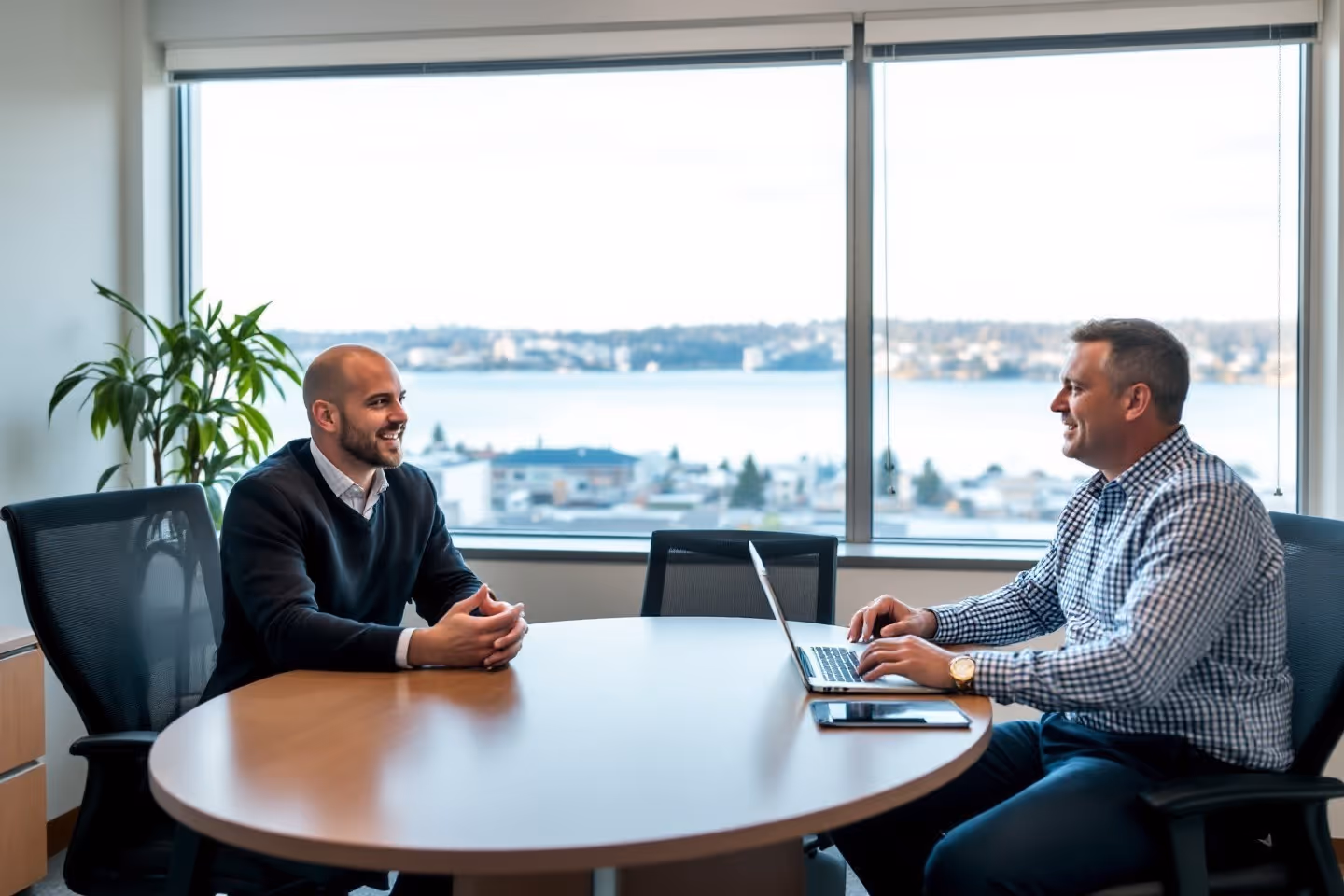 Two men smiling and talking across a round office table with a laptop in front of one, large window with cityscape and water view behind them.