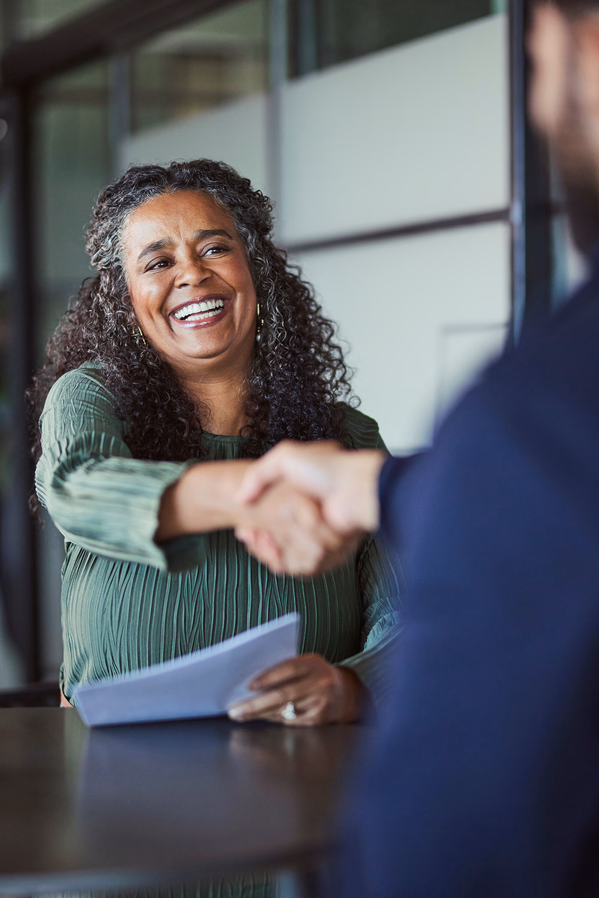 Smiling woman in green shirt shaking hands across a table holding papers during a meeting.