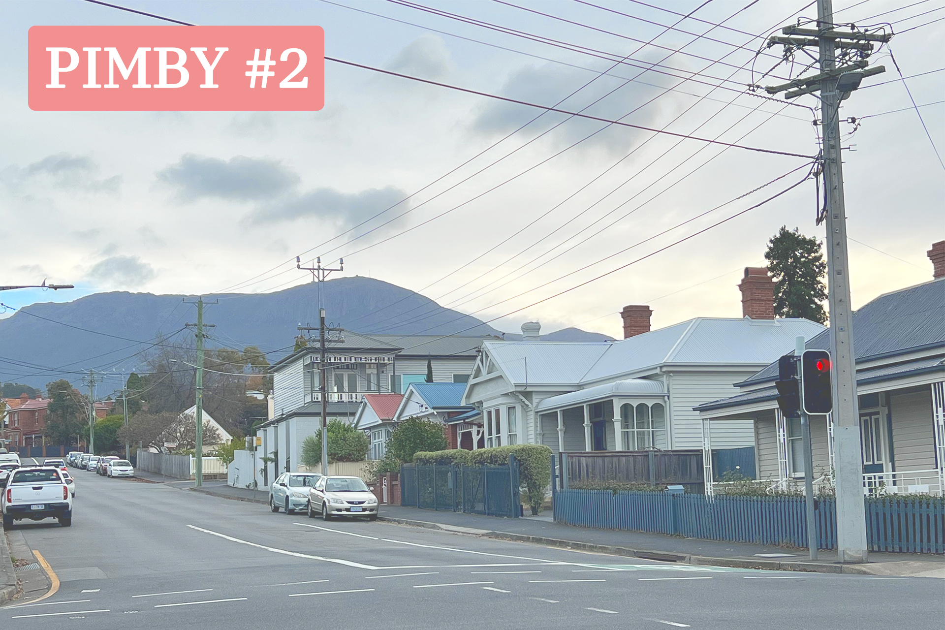 A Tasmanian street with several houses and a large sealed road. There are several cars parked. The sky has a few clouds and looks close to sunset. Mount Wellington is featured behind the houses.