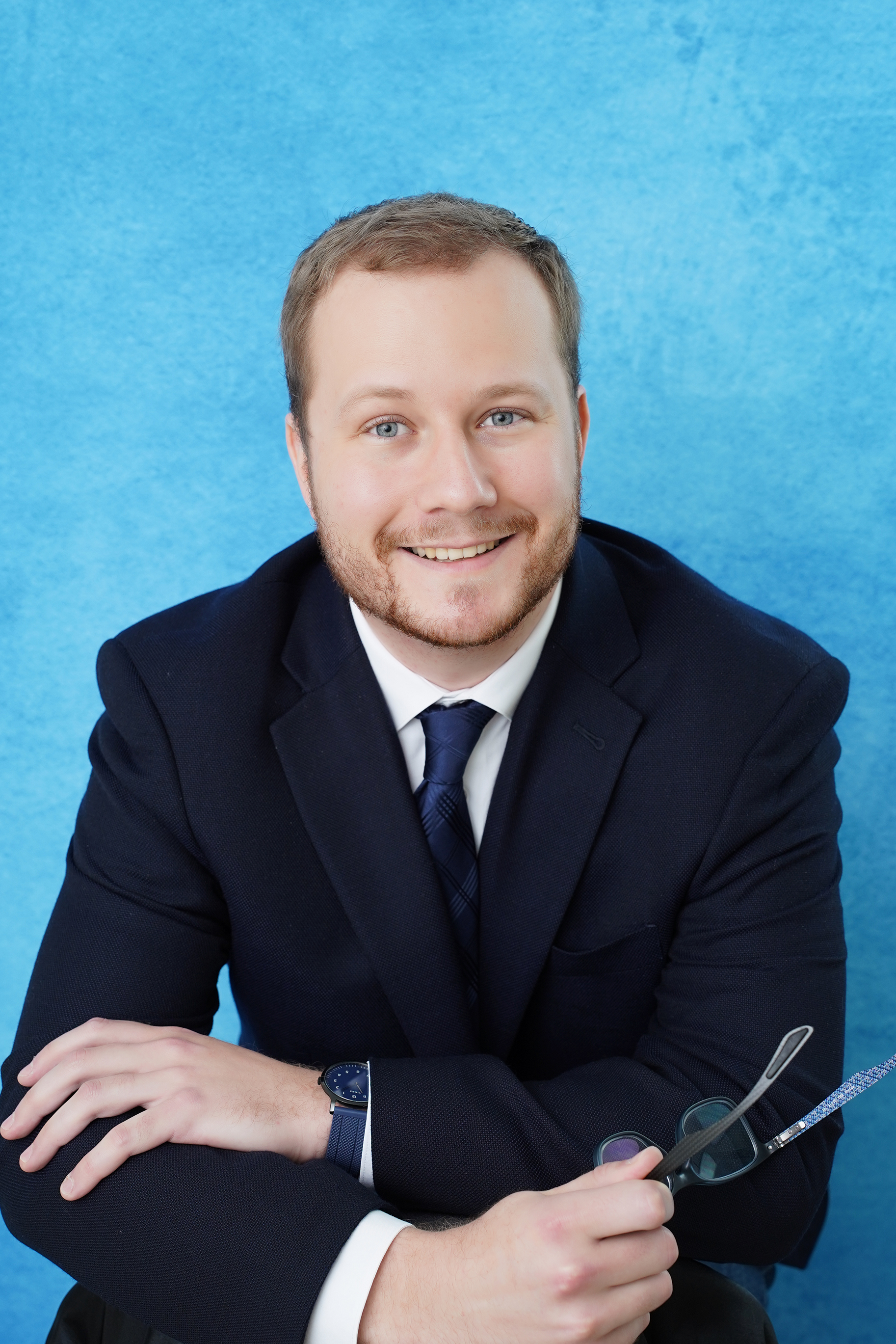 Smiling man in a navy suit and tie, holding glasses with a blue patterned temple against a bright blue background.
