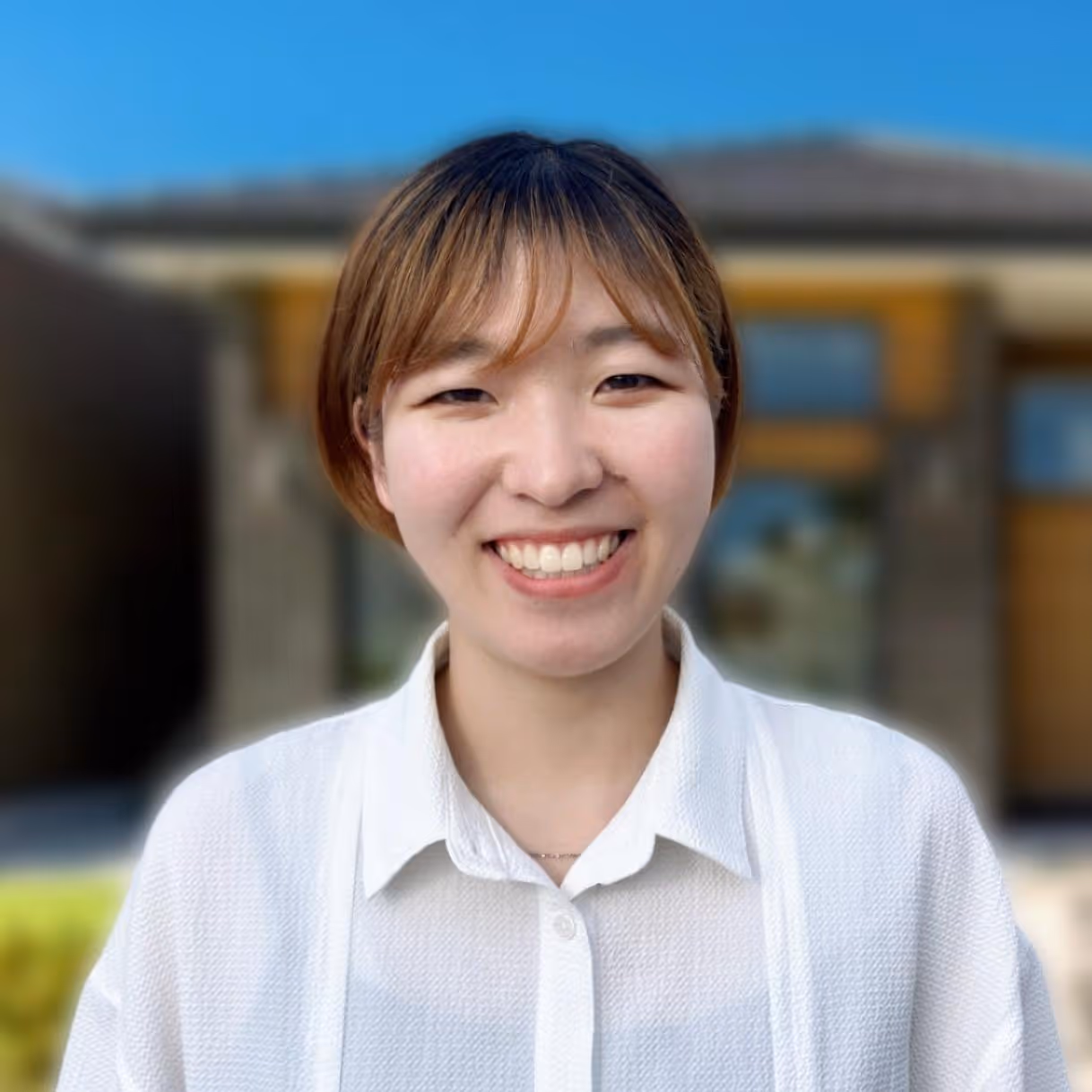 Smiling young woman with short brown hair wearing a white shirt outdoors in front of a building.