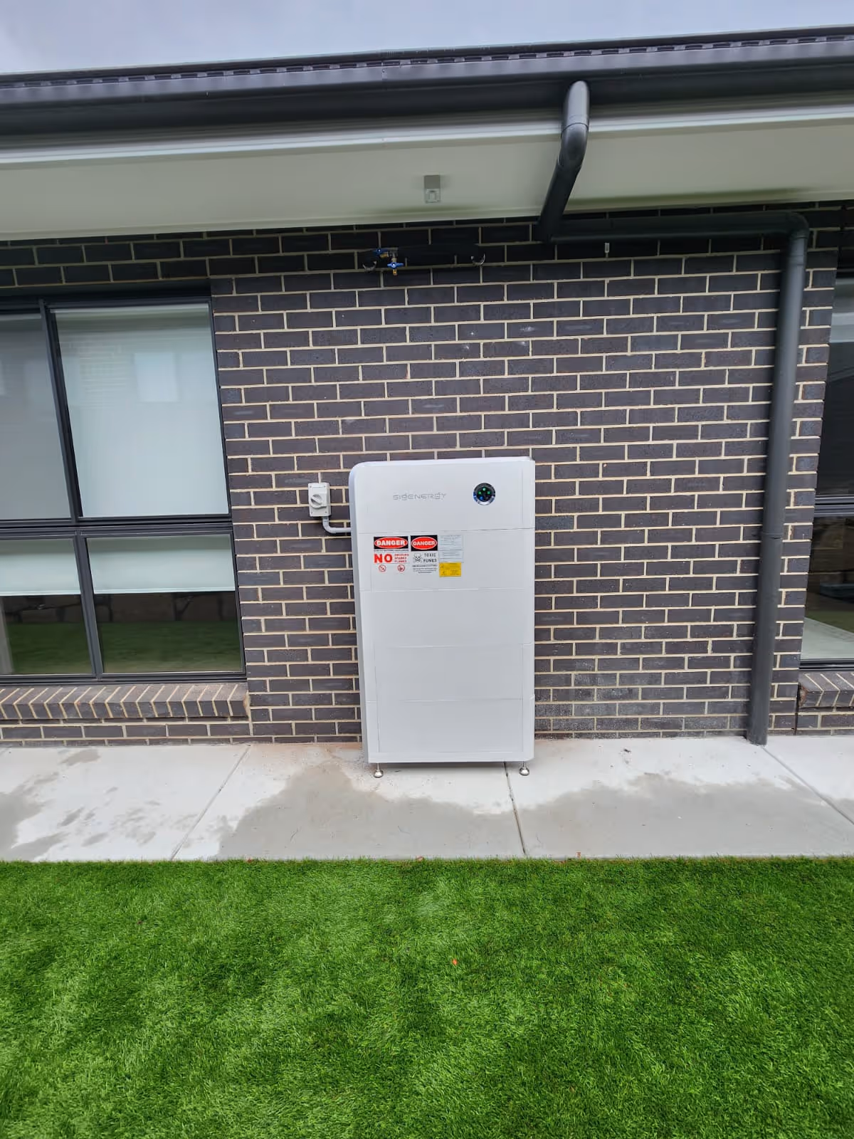 SigEnergy Battery (BESS) Home Energy Solution mounted on brick wall beside modern building with windows