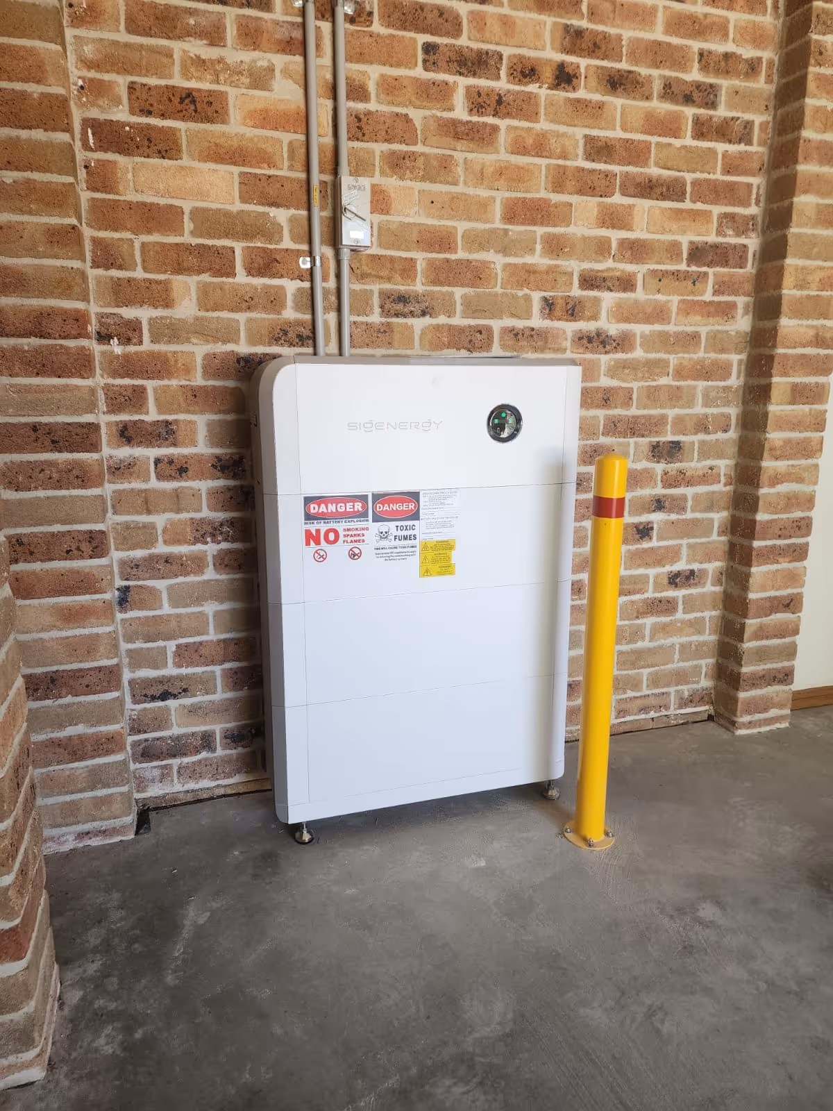 White SigEnergy Battery System (BESS) mounted on brick wall with danger labels and yellow protective post nearby