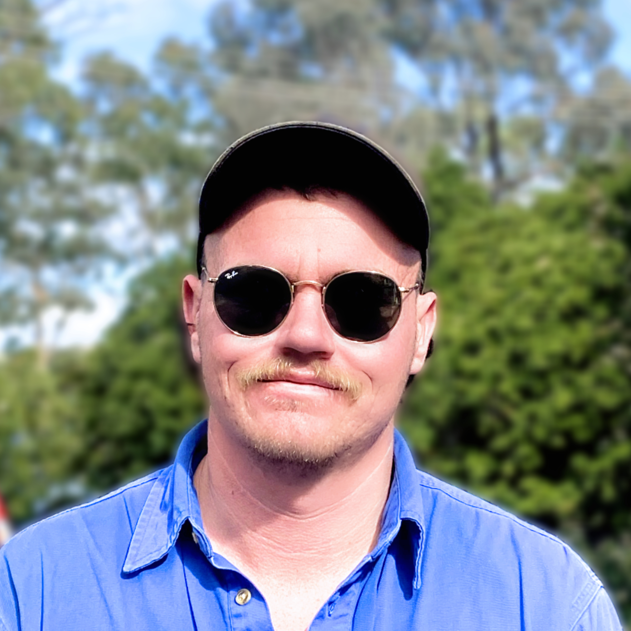 Man with a mustache wearing round sunglasses, a black cap, and a blue shirt outdoors.