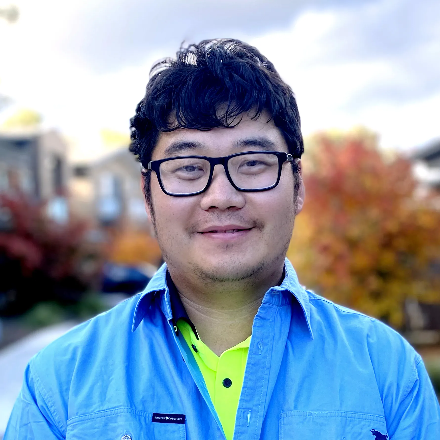 Smiling man with glasses wearing a blue shirt outdoors with autumn trees blurred in the background.