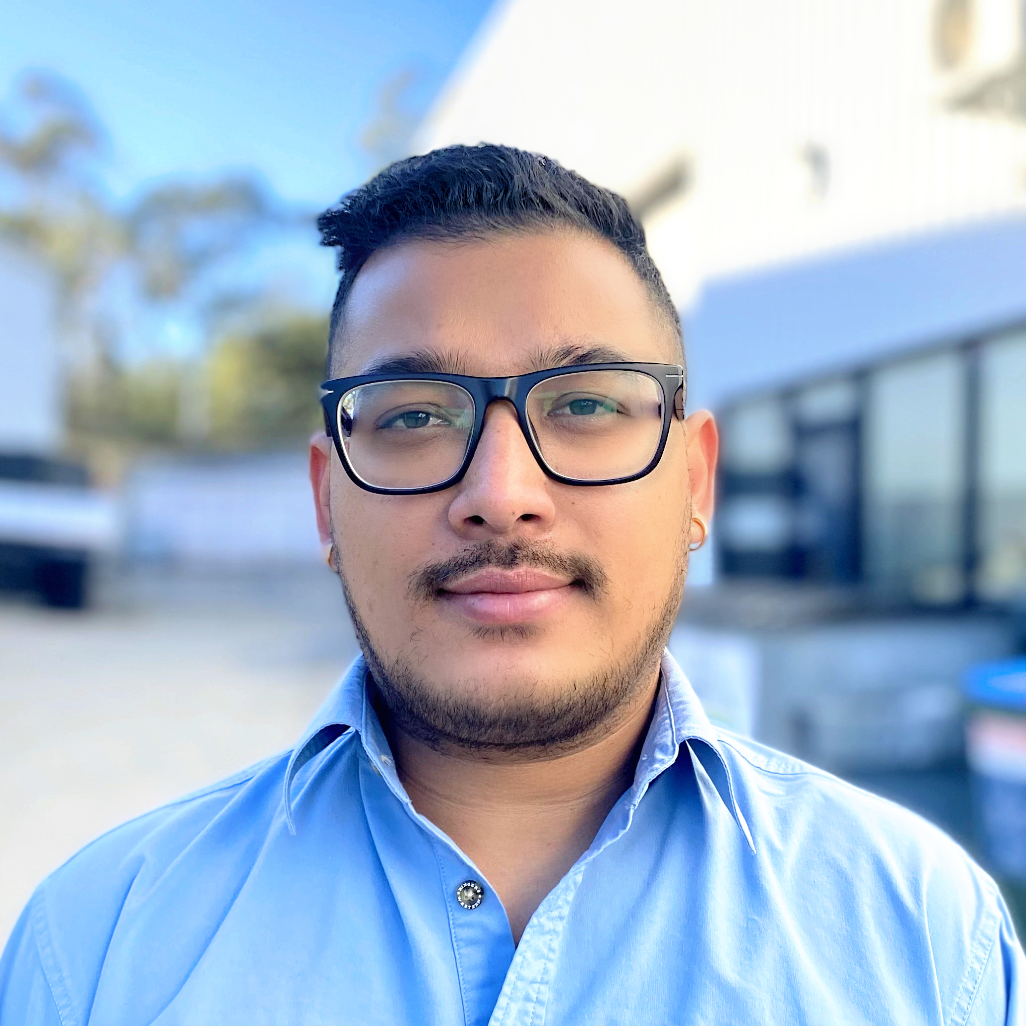 Man with short hair, black glasses, and blue shirt standing outdoors with blurred background.