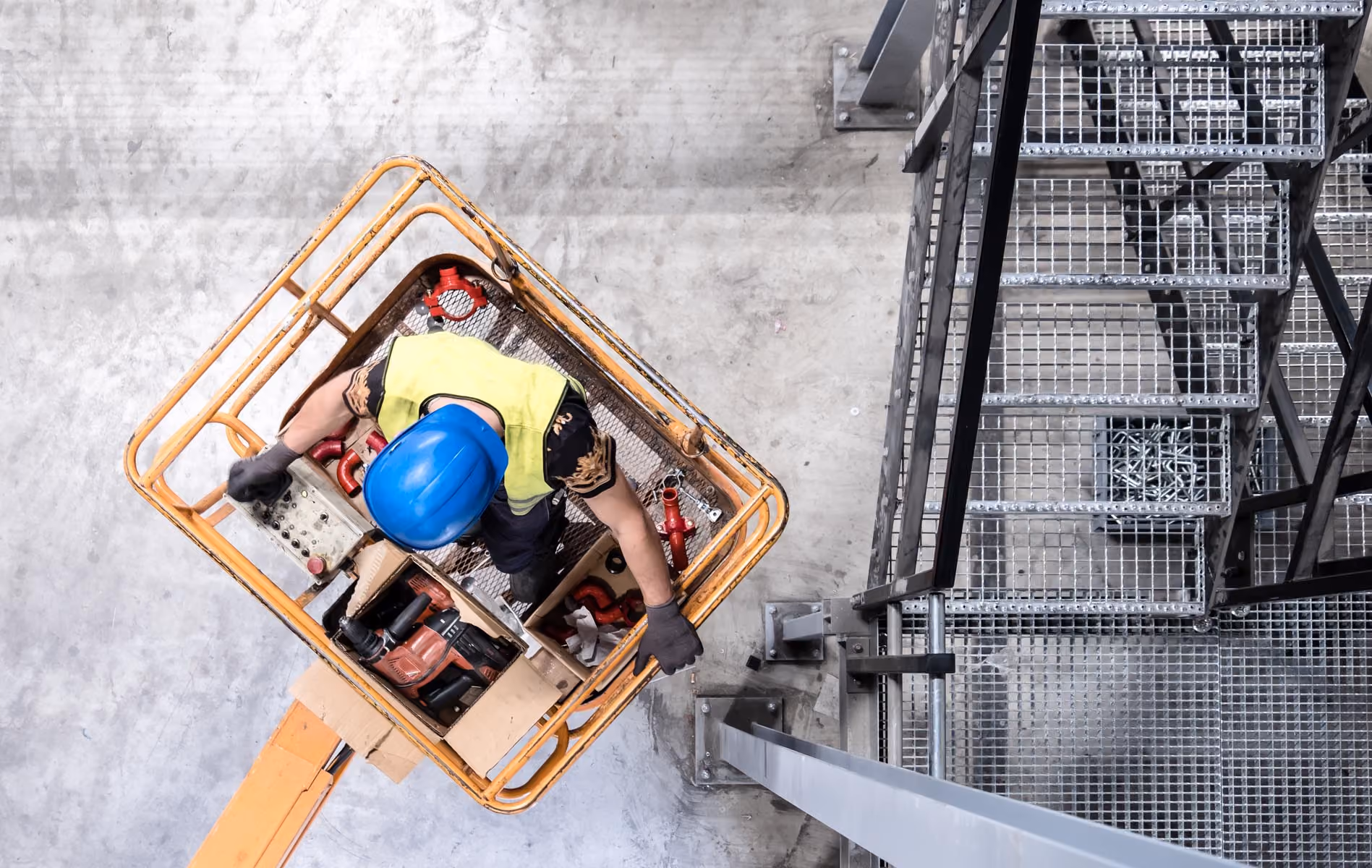 Worker in blue helmet and yellow vest on a yellow lift platform near a metal staircase indoors.